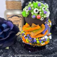 Cupcakes topped with multi-colored frosting (orange, blue, and green) and sprinkles and an Oreo dipped in chocolate and covered in green icing and a variety of sprinkles including eyeballs, bats, and ghosts to resemble a bubbling cauldron. Halloween decor surrounds the cupcakes.