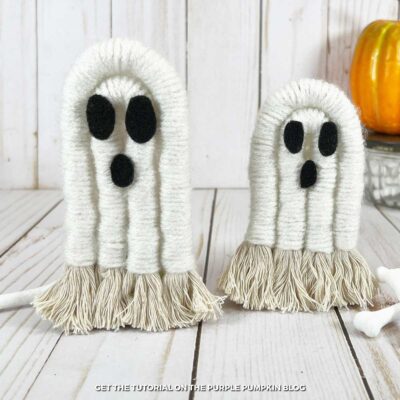 Rope that has been wrapped in white yarn, the ends frayed, and black felt eyes and mouth. Plastic pumpkin and bones are nearby as decoration.