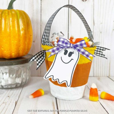 Two paper buckets, painted with yellow, orange, and white paint to look like candy corn, with gingham ribbon hands and cute ghost tags. Filled with paper filling and candy corns. Candy corns, candy bones, and a mini pumpkin surround.