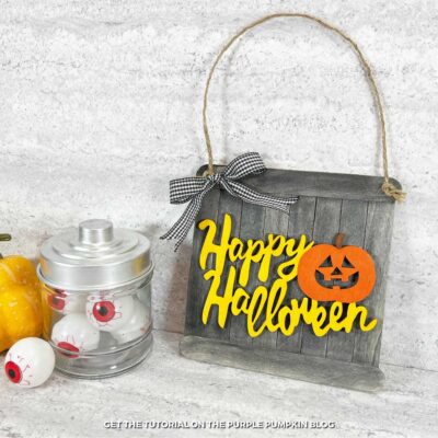 A sign made with craft sticks and decorated with the words Happy Halloween and Jack-o'lantern and a ribbon bow. A jar of fake eyeballs and mini pumpkin are sat beside it.
