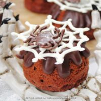 Mini chocolate bundt cakes topped with chocolate ganache and decorated with a chocolate spider and web. Halloween decorations are in the background.