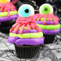 Cupcakes topped with layers of alternating purple and green frosting, with pink icing dripped on top to resemble blood and finished off with a candy eyeball. Halloween decorations are in the background.