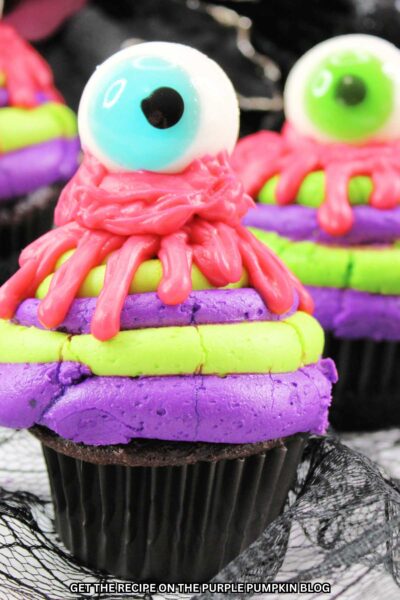 Cupcakes topped with layers of alternating purple and green frosting, with pink icing dripped on top to resemble blood and finished off with a candy eyeball. Halloween decorations are in the background.