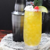 Glass of yellow cocktail with salt rim and garnished lime slice, cherry and a pineapple leaf. Cocktail shaker in the background.