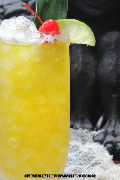 Close up of glass of yellow cocktail with salt rim and garnished lime slice, cherry and a pineapple leaf.