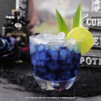 A rocks glass of deep blue cocktail garnished with lime slice and pineapple leaves. Halloween decor is in the background.