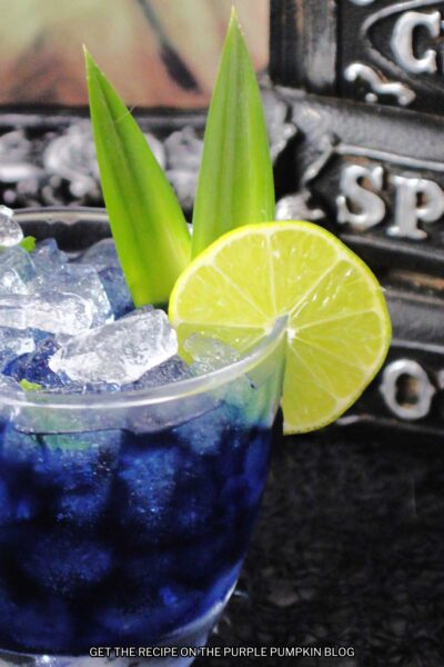 A rocks glass of deep blue cocktail garnished with lime slice and pineapple leaves. Halloween decor is in the background.