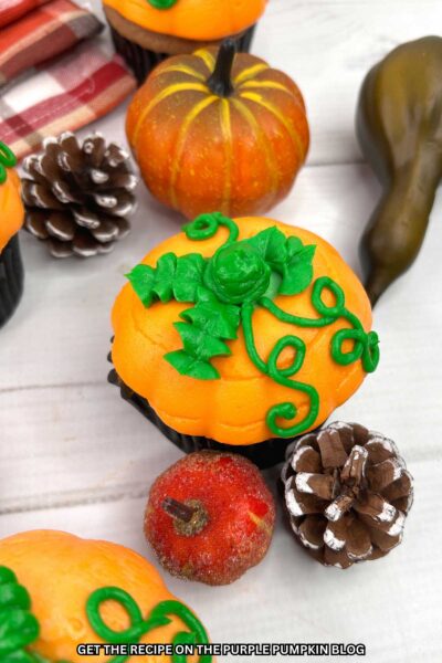 Cupcake topped with a mound of orange frosting to resemble a pumpkin, with green leaves and vines piped on top. Fall decor in the background.