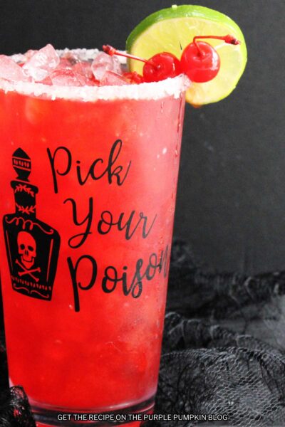 Red drink inside a glass that is embossed with a poison bottle and the words "Pick Your Poison". Glass is garnished with salt rim, red maraschino cherries and a slice of lime. Halloween decor is in the background.