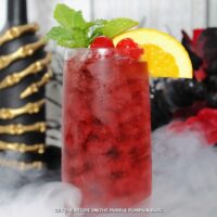 A glass of deep red cocktail garnished with cocktail cherries, slice of orange, and mint leaves. Halloween decorations are in the background.