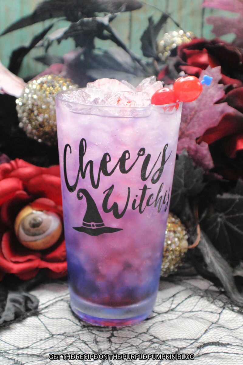 A glasses embossed with "Cheers Witches" and a witch hat, filled with a pink-purple drink and garnished with cocktail cherries. Halloween decorations are in the background.