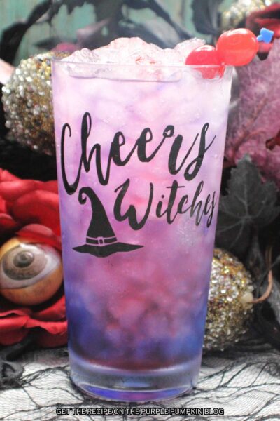 A glasses embossed with "Cheers Witches" and a witch hat, filled with a pink-purple drink and garnished with cocktail cherries. Halloween decorations are in the background.