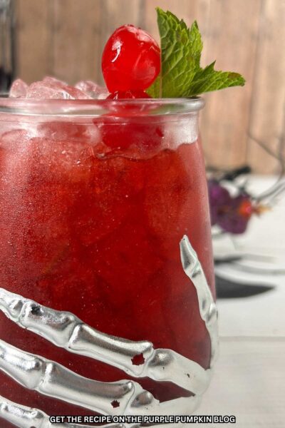 Deep red cocktail in a glass adorned with a skeleton hand and garnished with a cocktail cherry and fresh mint leaves. Halloween decorations are in the background.