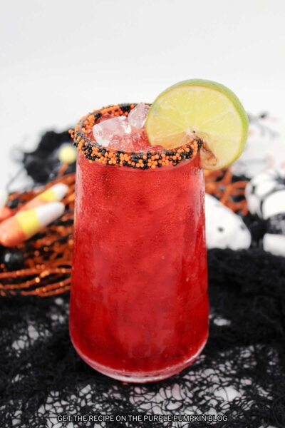 Cocktail glass filled with red drink, rim covered in orange and black sprinkles, and garnished lime slice. Halloween decorations are in the background.