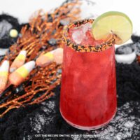 Cocktail glass filled with red drink, rim covered in orange and black sprinkles, and garnished lime slice. Halloween decorations are in the background.