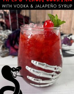 Deep red cocktail in a glass adorned with a skeleton hand and garnished with a cocktail cherry and fresh mint leaves. Halloween decorations are in the background. Text overlay says: Viper's Venom Cocktail with Vodka and Jalapeno Syrup