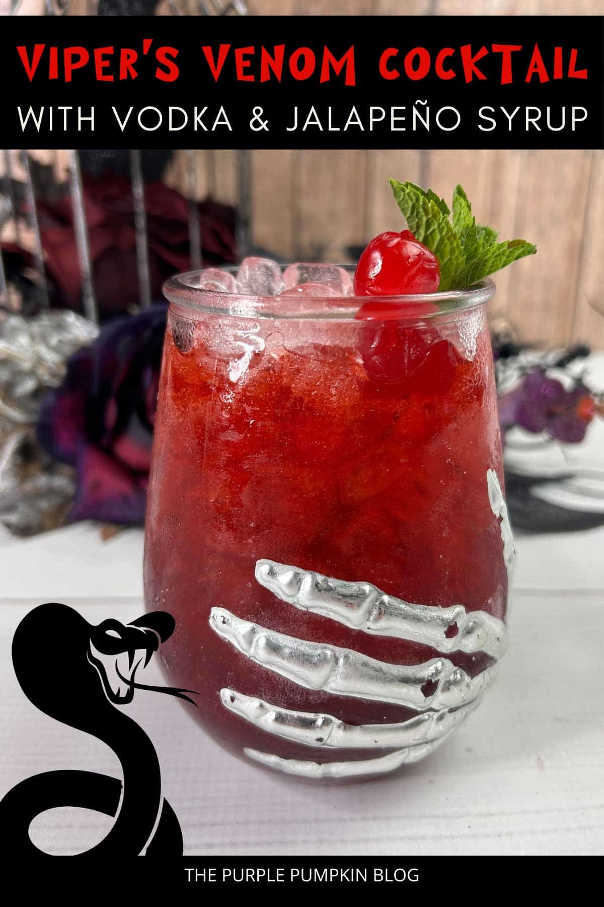Deep red cocktail in a glass adorned with a skeleton hand and garnished with a cocktail cherry and fresh mint leaves. Halloween decorations are in the background. Text overlay says: Viper's Venom Cocktail with Vodka and Jalapeno Syrup