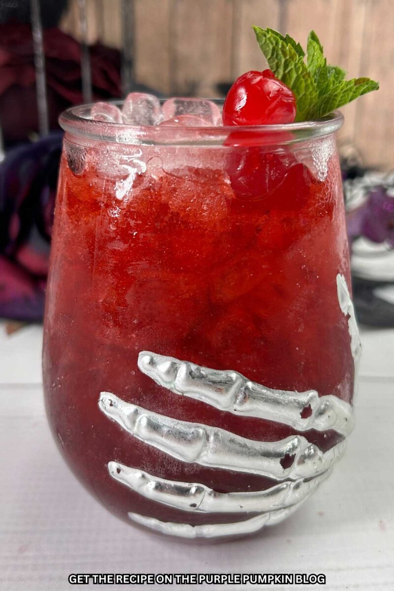 Deep red cocktail in a glass adorned with a skeleton hand and garnished with a cocktail cherry and fresh mint leaves. Halloween decorations are in the background.