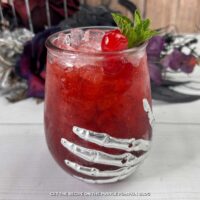Deep red cocktail in a glass adorned with a skeleton hand and garnished with a cocktail cherry and fresh mint leaves. Halloween decorations are in the background.