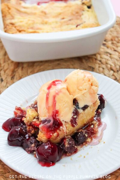 A plate with a portion of cherry dump cake topped with additional cherries and a scoop of ice cream. The dish of cake is in the background.