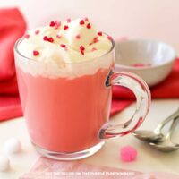 A glass mug filled with light red colored hot chocolate topped with whipped cream and heart-shaped sprinkles.