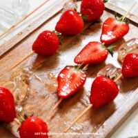 Skewered fresh strawberries that have been dipped in sugar syrup. Placed on a wooden board and surrounded by a few ice cubes.