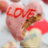 A heart-shaped cake pop covered with pink chocolate the word LOVE piped on. The pop has a bite taken out of it