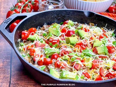 Side angle of a cast iron skillet filled with a colorful taco skillet made with ground beef, shredded cheese, avocado chunks, and diced tomatoes. The dish is garnished with green onions and set on a wooden table. In the background, there's a jar of taco seasoning, fresh cherry tomatoes on the vine, a bowl of tortilla chips, and a red-striped napkin for a cozy presentation.