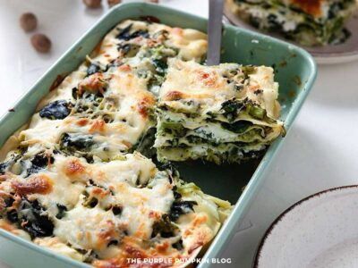 Baking dish filled with freshly baked vegetarian lasagna featuring layers of spinach, broccoli, ricotta, and béchamel sauce, with one slice being lifted to reveal creamy green vegetable layers. Melted mozzarella and golden cheese top the dish, with a white plate ready to serve in the foreground.