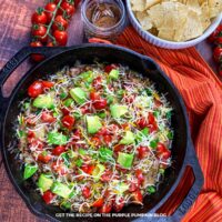Cast iron skillet filled with a colorful taco skillet made with ground beef, melted shredded Mexican cheese blend, diced tomatoes, avocado chunks, and sliced green onions. The pan sits on a rustic wooden table next to a bowl of tortilla chips, a jar of taco seasoning, and a vine of fresh cherry tomatoes. A red and orange striped cloth adds a pop of color. Overhead view with vibrant, appetizing presentation.