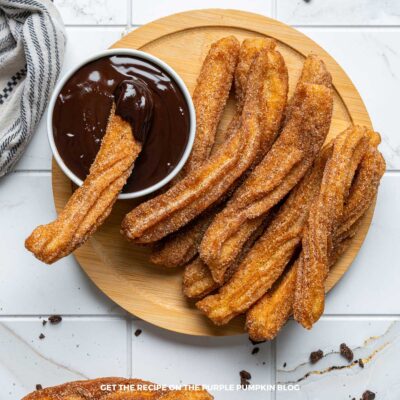 A round wooden board filled with crispy Mexican churros and a bowl of rich chocolate dipping sauce, one churro partially dipped.