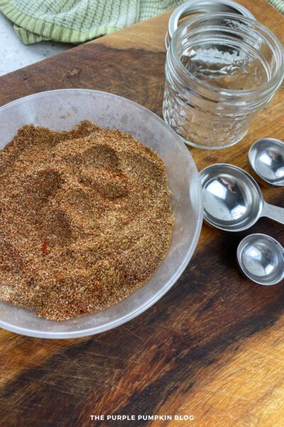 Overhead view of a bowl filled with a blended homemade taco seasoning mixture featuring a warm-toned combination of spices. Next to the bowl are a glass jar with lid and a set of metal measuring spoons on a rustic wooden cutting board. A green kitchen towel is visible in the background. The image suggests storing the blend in an airtight container.
