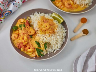 Overhead shot of a plate of Shrimp Tikka Masala served with white basmati rice, garnished with fresh cilantro and a wedge of lime. The shrimp are coated in a creamy, orange-hued curry sauce made with onions, tomatoes, and Indian spices. Two wooden measuring spoons with ground turmeric and garam masala are placed nearby, and a colorful napkin adds detail in the top left.
