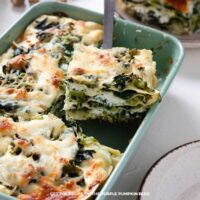 A close-up of a freshly baked vegetarian lasagna in a light green baking dish, layered with spinach, broccoli, ricotta, mozzarella, and béchamel sauce. A serving spatula lifts a neatly cut square of the lasagna to reveal the creamy green vegetable layers.