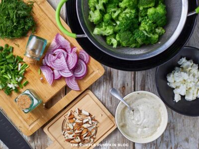 Salad bowl filled with broccoli, red onion, dill, chives, and almonds, ready to be tossed; surrounding ingredients and chopped vegetables visible on the wooden surface.