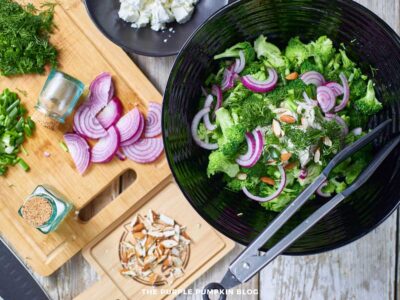 Chopped red onion, herbs, and almonds on a cutting board next to a bowl of creamy dressing, crumbled feta, and blanched broccoli in a bowl.
