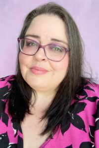 Michelle from The Purple Pumpkin Blog, smiling warmly at the camera. She has long dark hair with natural silver streaks, purple glasses, and soft pink makeup. She’s wearing a bold pink floral top, with a gentle pastel purple background that adds a whimsical, welcoming vibe.