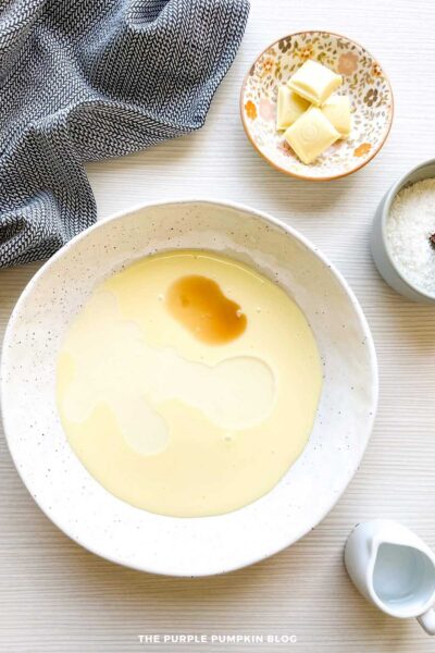 Bowl of sweetened condensed milk with tequila and vanilla extract just added; ingredients are resting on the surface before mixing.