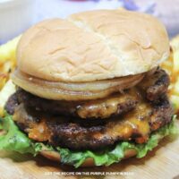Close view of smash burger with two beef patties, grilled onions, cheddar cheese, lettuce, and a side of fries.