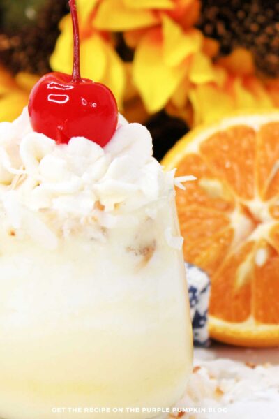 Dole whip shot with creamy layers, shredded coconut, and cherry on top with orange slices in background