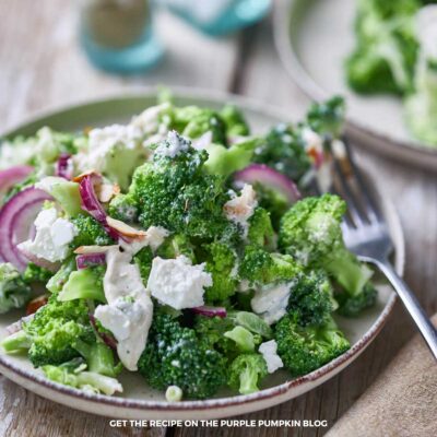 Broccoli salad on a rustic plate with visible crumbled feta, sliced red onions, and a creamy dressing, served with a fork on a wooden table.