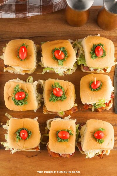 A set of BLT sliders arranged on a wooden board, each topped with a cherry tomato and a piece of kale secured by a toothpick.