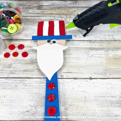 Uncle Sam wooden spoon craft laid out on wood background with buttons, glue gun, and craft supplies.
