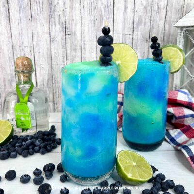 Shot of two vibrant frozen blueberry limeade margaritas with lime and blueberry garnish, set beside a tequila bottle and checkered cloth.