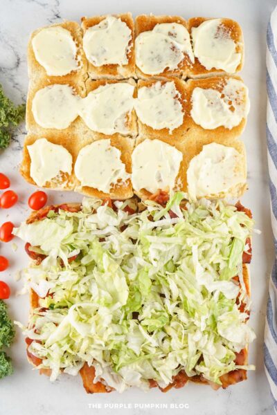 The bottom halves of the slider buns fully layered with shredded lettuce, with mayo-coated top buns positioned above.