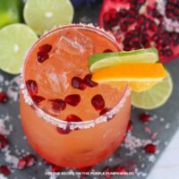 Overhead view of pomegranate orange margarita with salted rim, ice, pomegranate seeds, lime slice, and orange slice garnish.