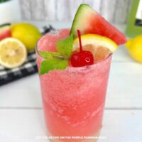 A tall glass of frozen watermelon lemonade garnished with a wedge of watermelon, lemon slice, cherry, and fresh mint sprig, no text overlay.