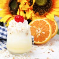 Close-up of a single Dole Whip shot topped with whipped cream and a cherry, with sliced oranges and sunflowers in the background.