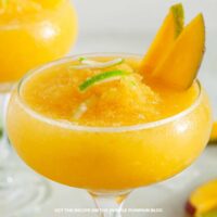 A close-up of a frozen Mango Coconut Daiquiri in a coupe glass, garnished with two mango slices and lime zest strips, no text overlay.