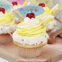 A single pina colada cupcake with tropical decorations including a cocktail umbrella, cherry, and pipette, surrounded by more cupcakes on a wooden board.